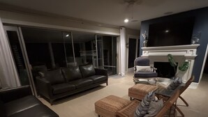 Een woonkamer met een zwarte bank, een houten stoel en een kleine tafel. Er is een open haard en een televisie bevestigd aan de muur.