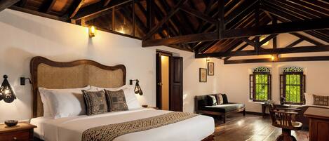 A spacious room with a large bed, wooden beams, a desk, and a view of greenery through windows.