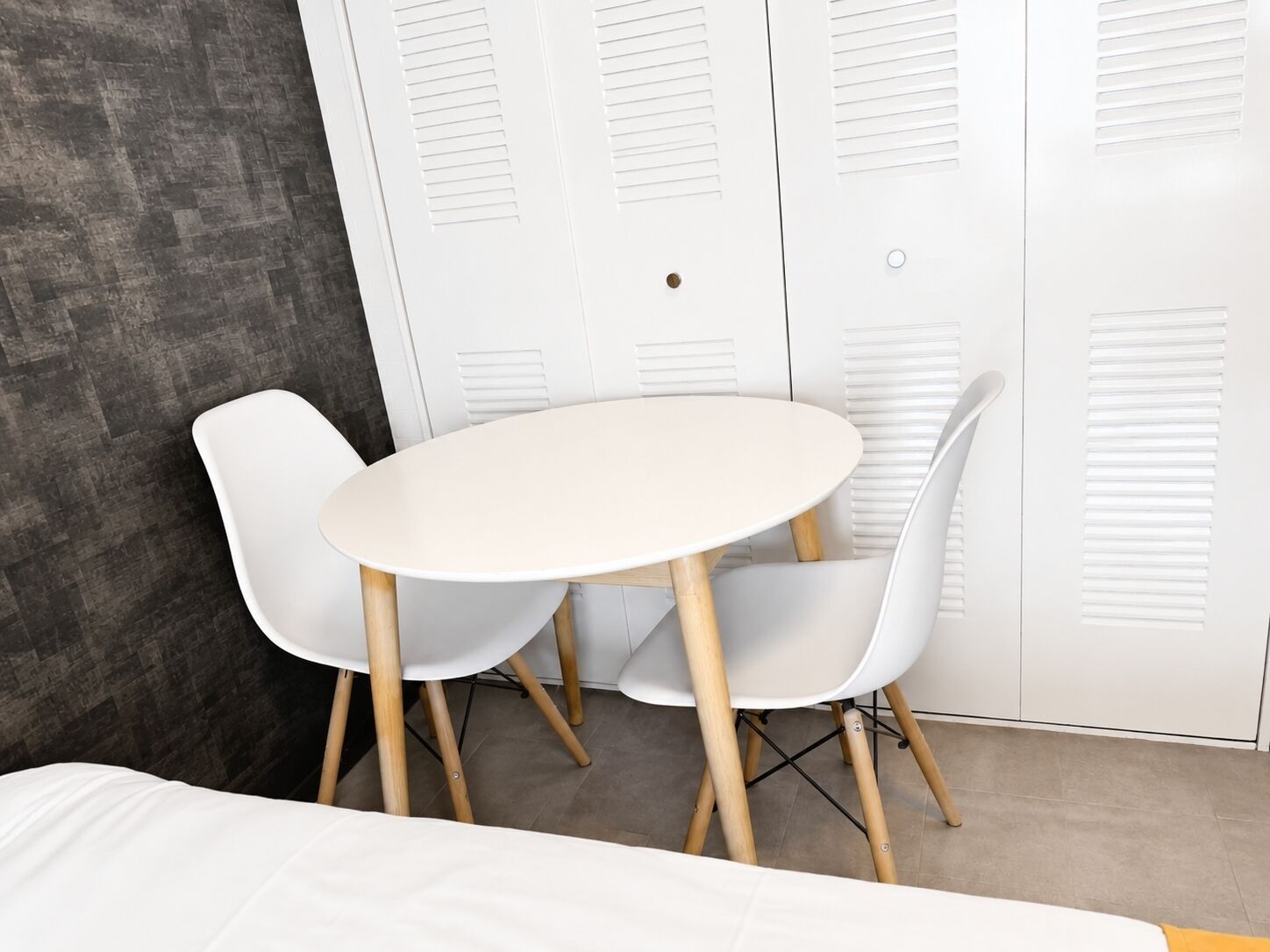 Una habitació amb un llit, una taula rodona blanca i dues cadires, i un armari amb portes a lamel·les.