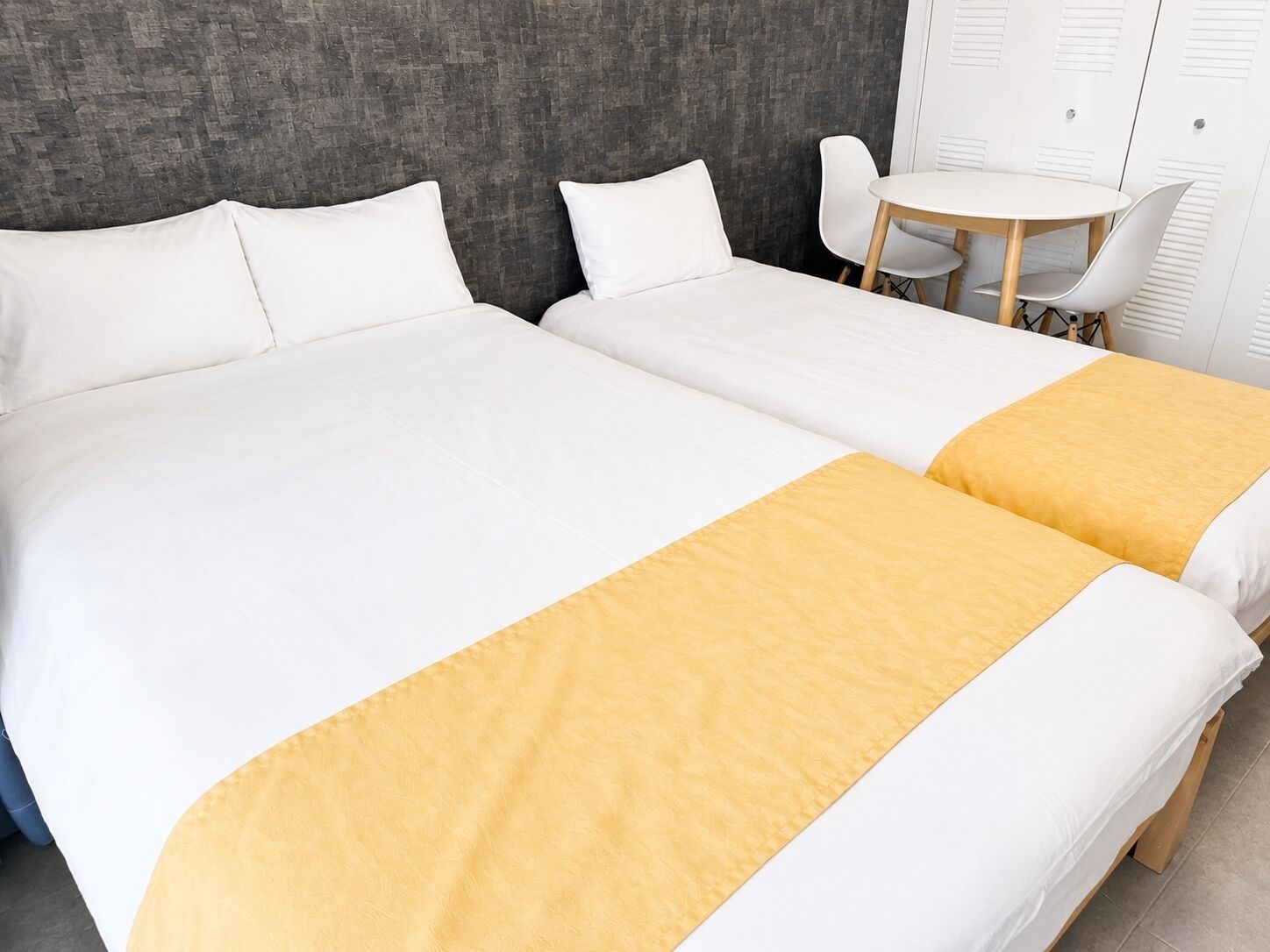 A double bed with white and yellow bedspreads, a round table with chairs, and a grey textured wall.