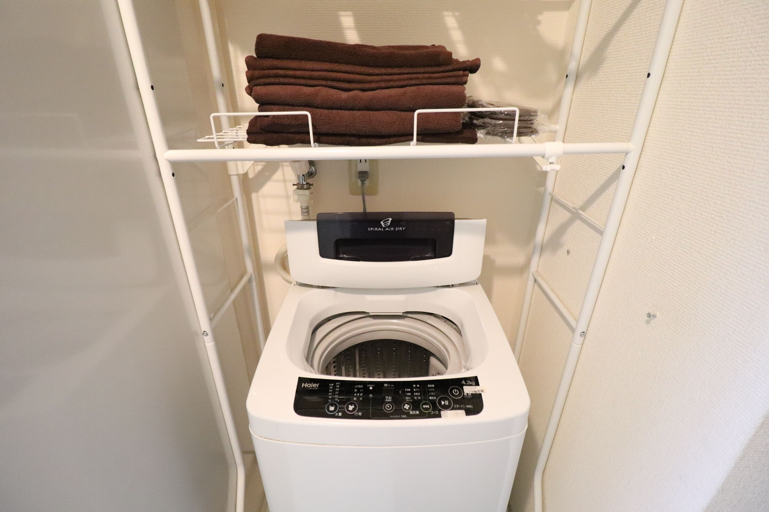 A compact, white, wall-mounted washer and dryer unit with a control panel, located in a small, enclosed laundry room with a shelf above holding folded towels.