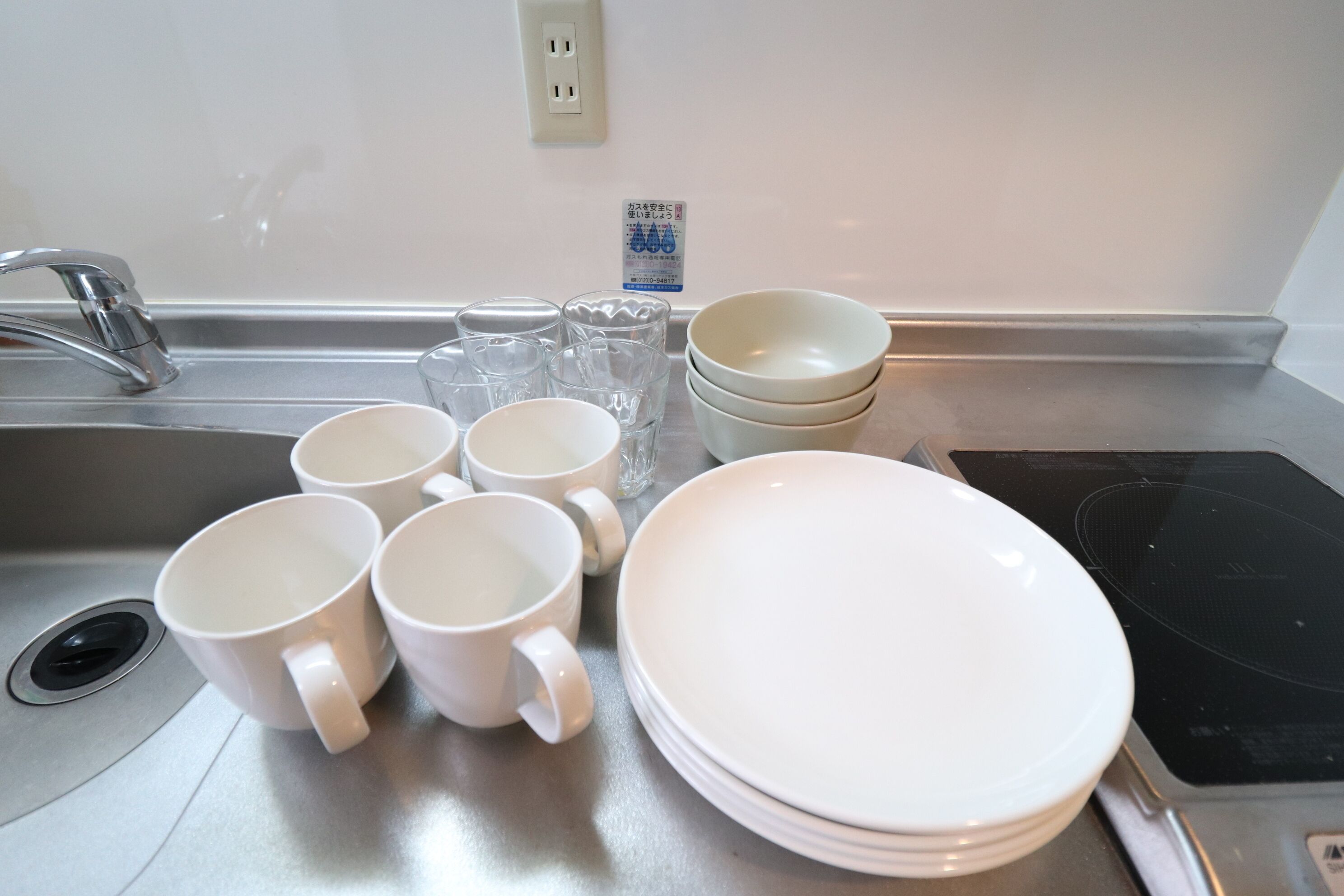 Un évier de cuisine avec des tasses blanches, des verres transparents et des assiettes blanches empilées sur le comptoir.