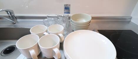 A kitchen sink with white mugs, clear glasses, and white plates stacked on the counter.