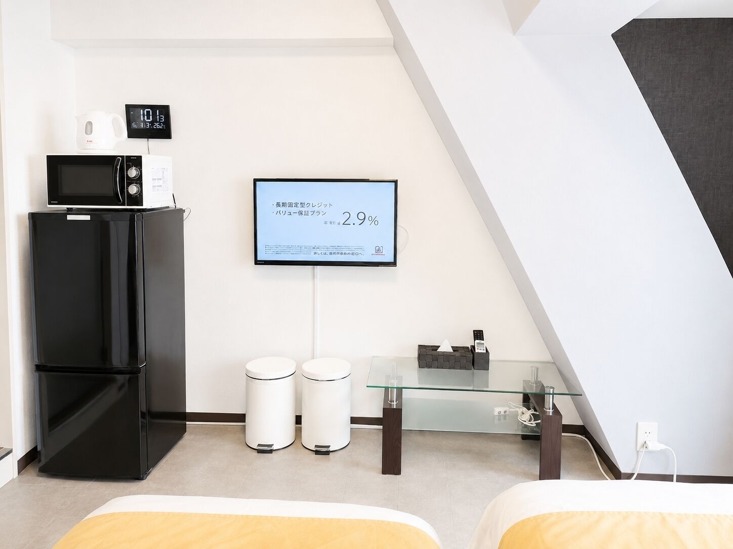 Une chambre d’hôtel moderne équipée d’un réfrigérateur noir, d’un four à micro-ondes, d’une horloge numérique, d’une table en verre avec deux poubelles blanches et d’un lit avec une tête de lit jaune.