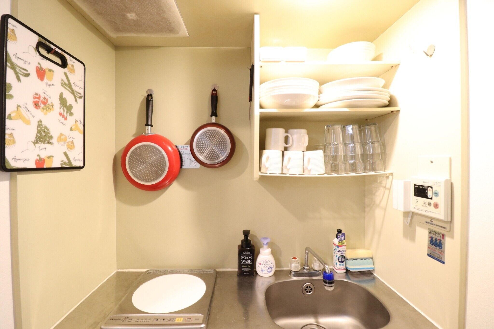 Una cocina con un lavamanos, una sartén roja, una sartén blanca y una estantería con platos y tazas.