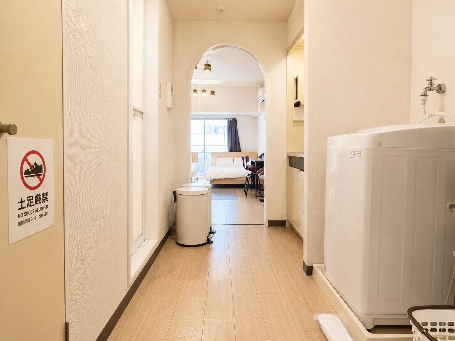 Een hotelgang met een bordje dat aangeeft dat schoenen niet zijn toegestaan, een prullenbak, een bed, een bureau en een wasmachine.