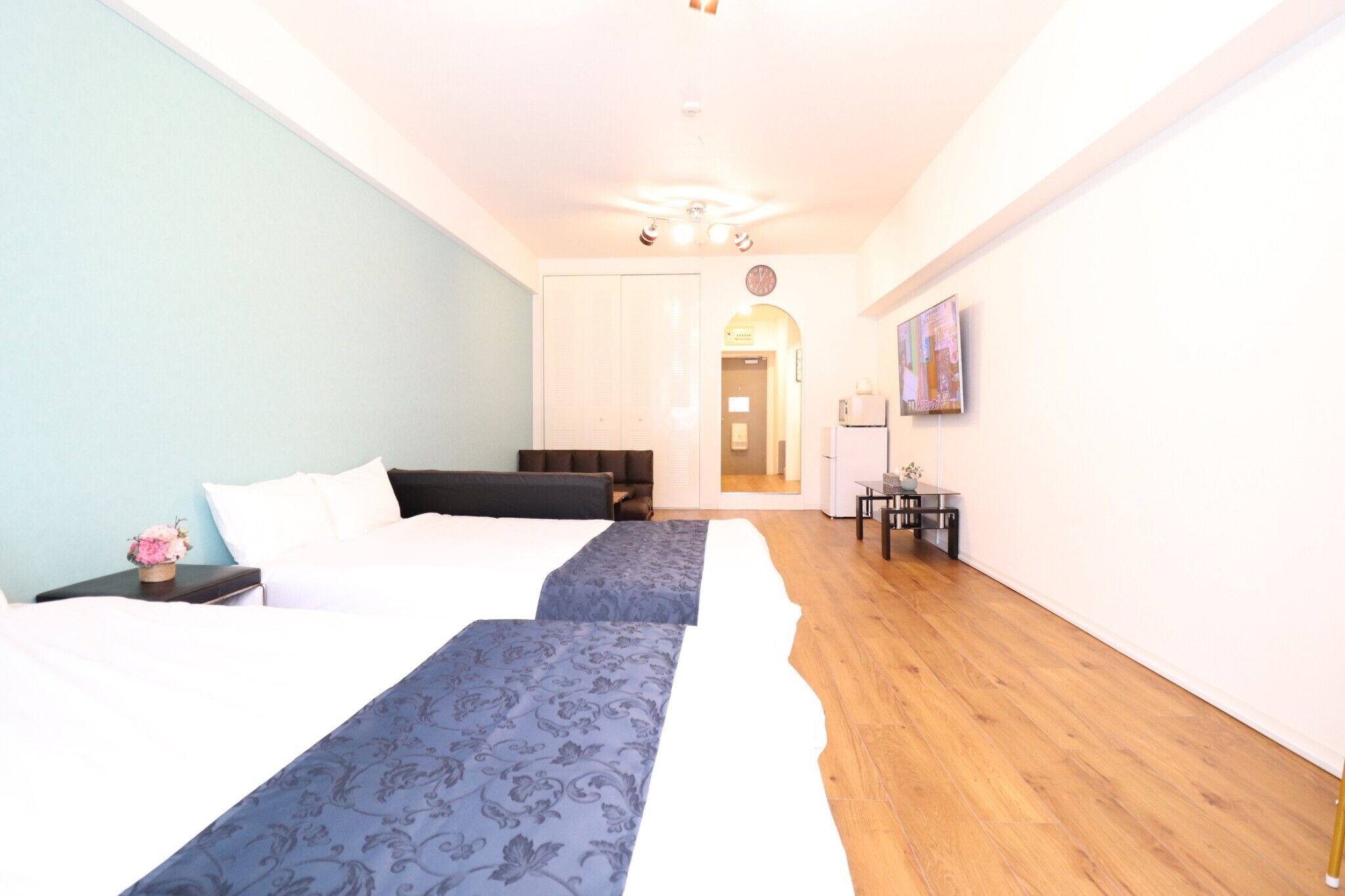 A hotel room with two beds, a wooden floor, a clock on the wall, and a TV on the right side.