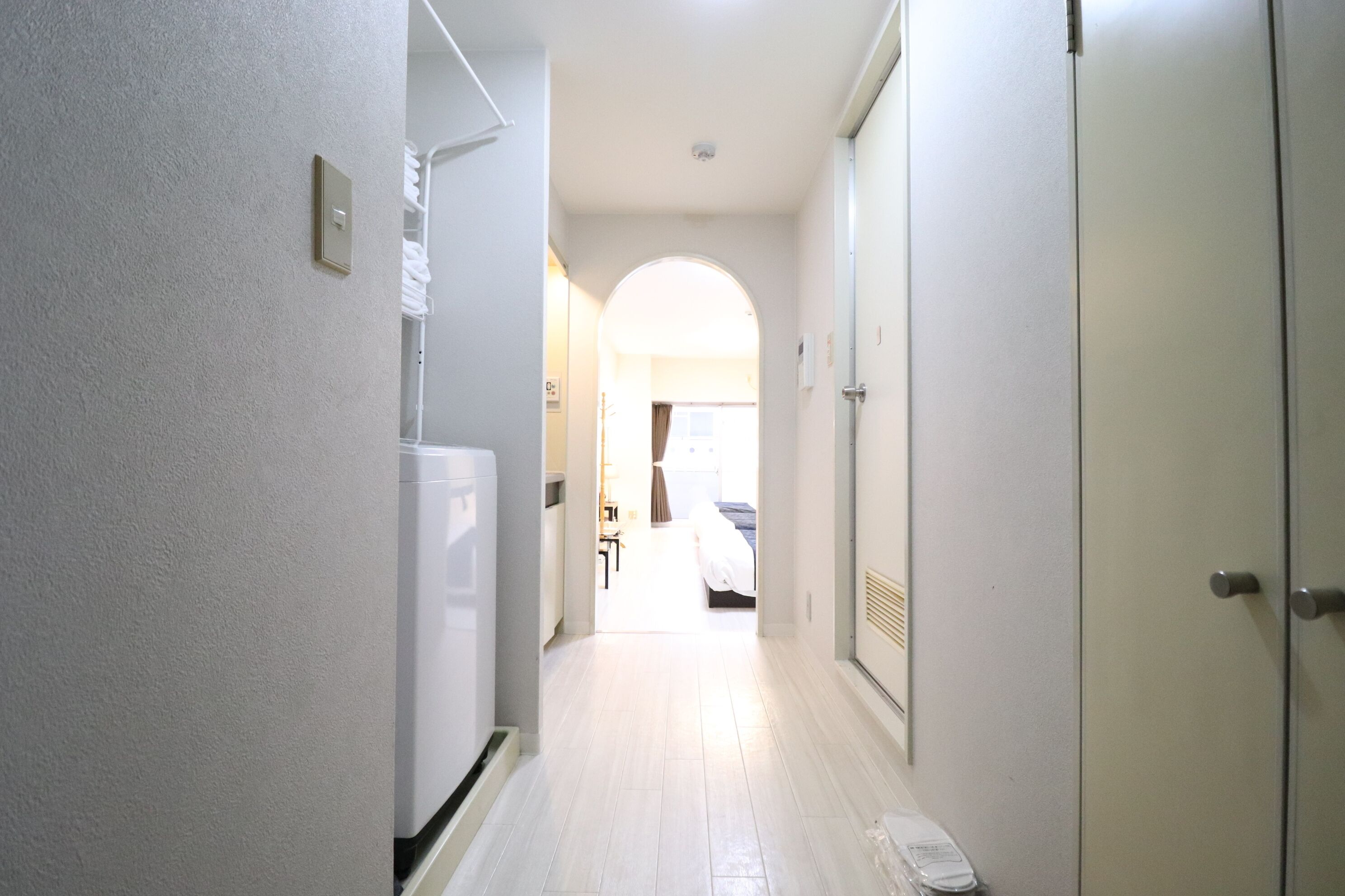 Un couloir étroit, avec un réfrigérateur blanc sur le côté gauche, une porte sur le côté droit et, au fond, une porte en arc qui mène à une pièce contenant un lit et une chaise.