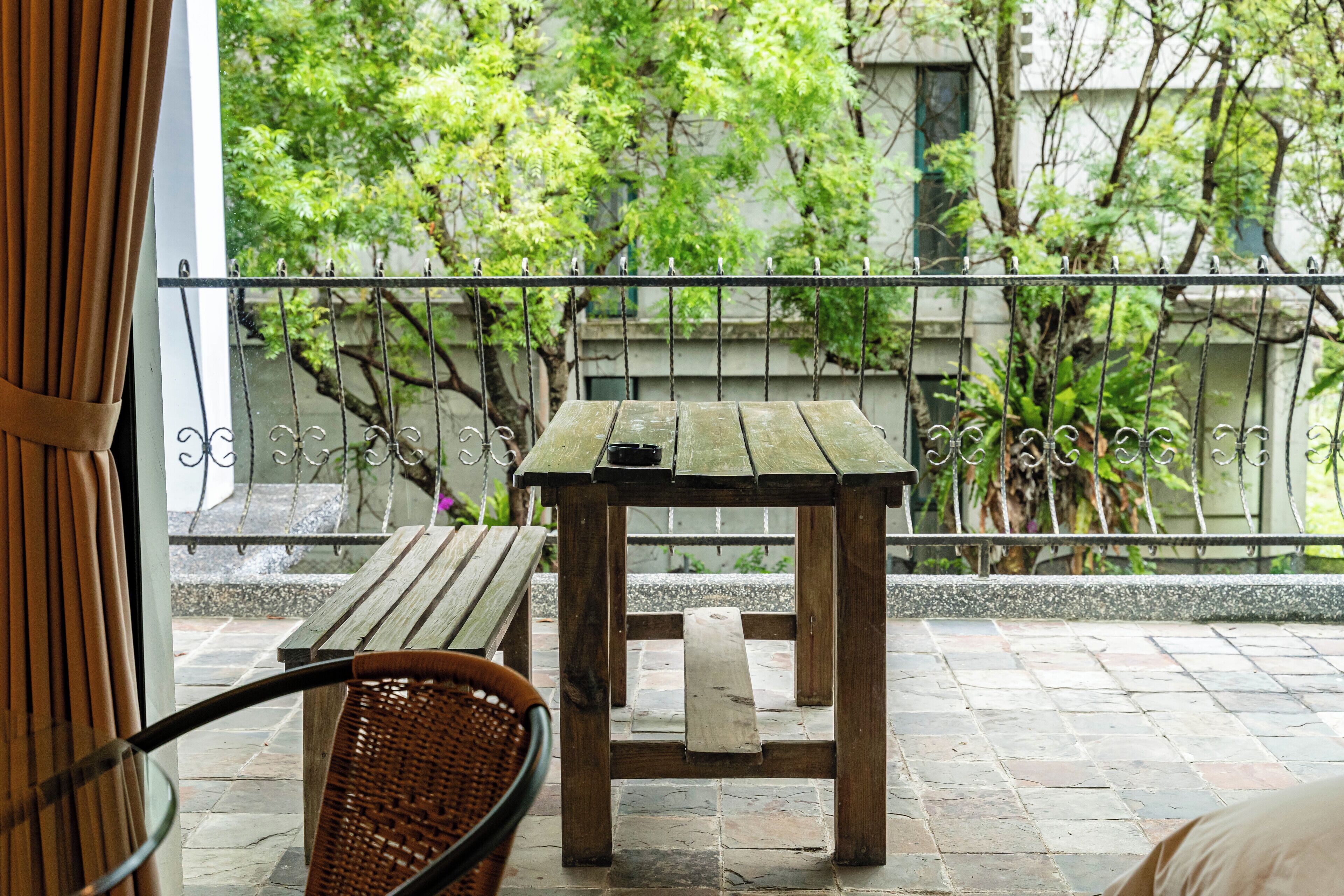 A wooden table and wicker chair on a balcony with a metal railing, stone tiles, and greenery in the background.