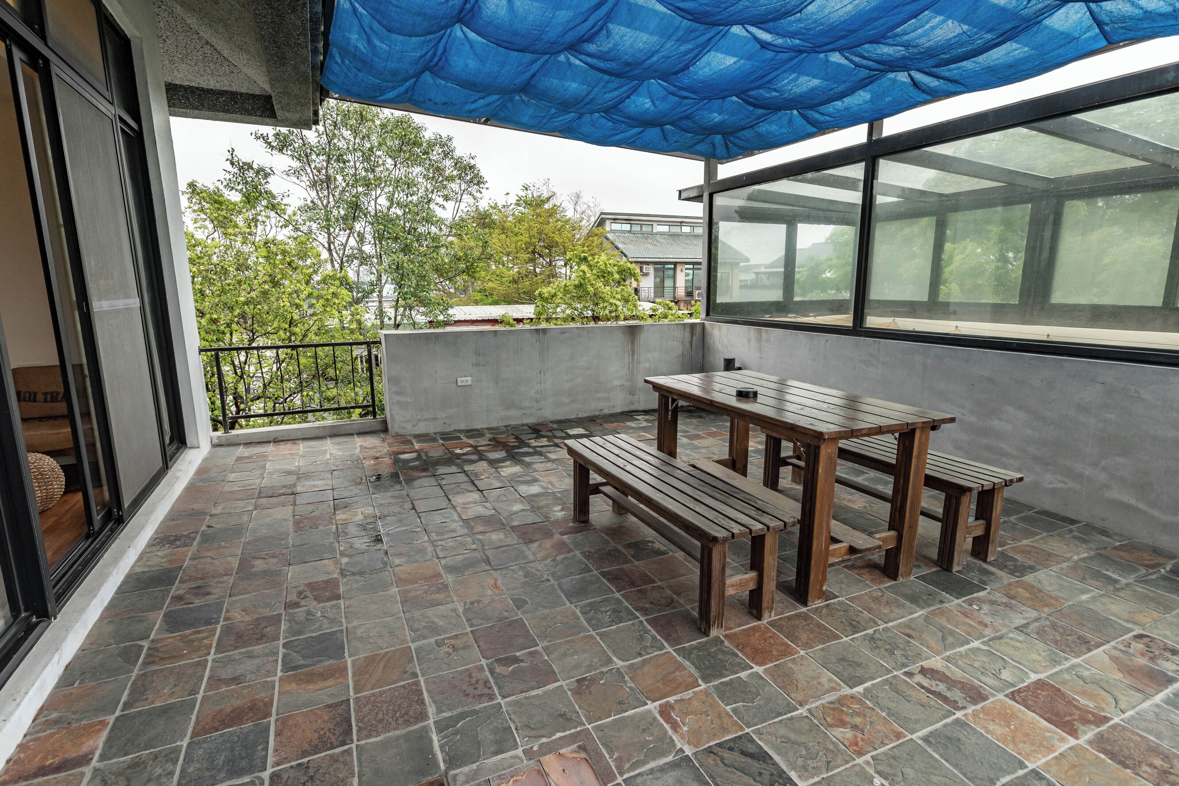 A covered outdoor area with a wooden table and benches, a view of trees and buildings in the background, and a concrete wall.