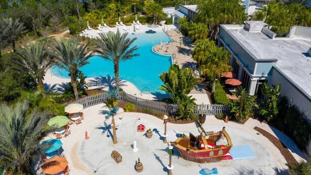 An aerial view of a resort with a central swimming pool, surrounded by palm trees, lounge chairs, and a small boat. - Game Room House - Resort Access (Davenport)