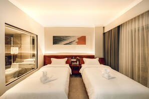 A hotel room with two beds, a nightstand, a painting, a window with a view of a bathroom, and a curtain.