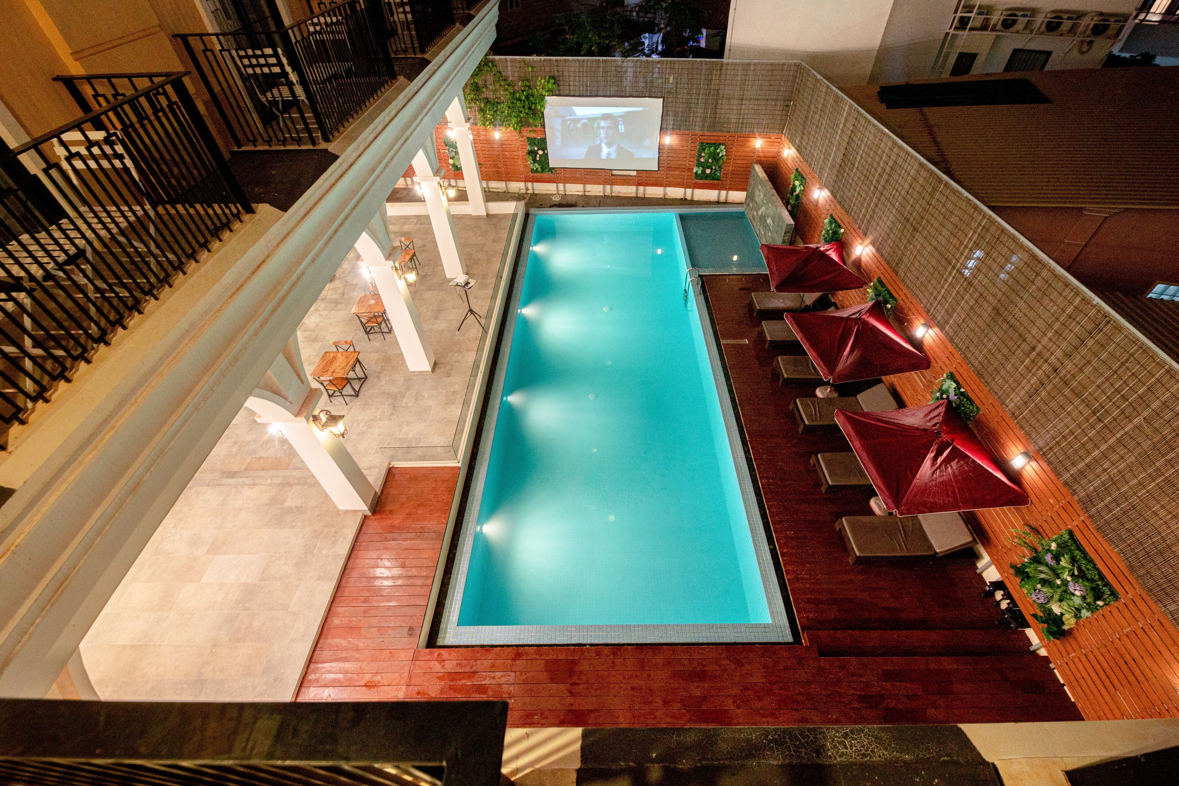 A rooftop swimming pool with a long, narrow shape, surrounded by wooden deck seating and red umbrellas.