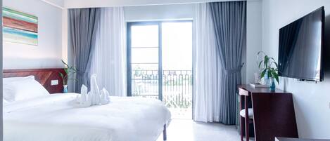 A hotel room with a large bed, a TV mounted on the wall, a balcony with curtains, and a plant on the side table.