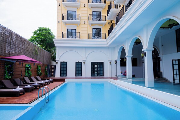 A swimming pool with lounge chairs and umbrellas, surrounded by a building with balconies and arched walkways.