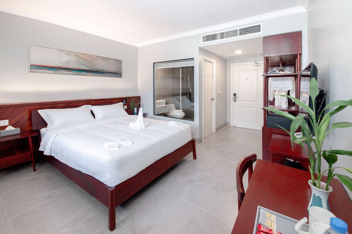 A hotel room with a bed, a desk, a chair, a plant, and a bathroom with a shower and toilet.