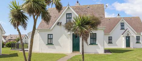A white building with a green door and windows, surrounded by a lawn and palm trees.
