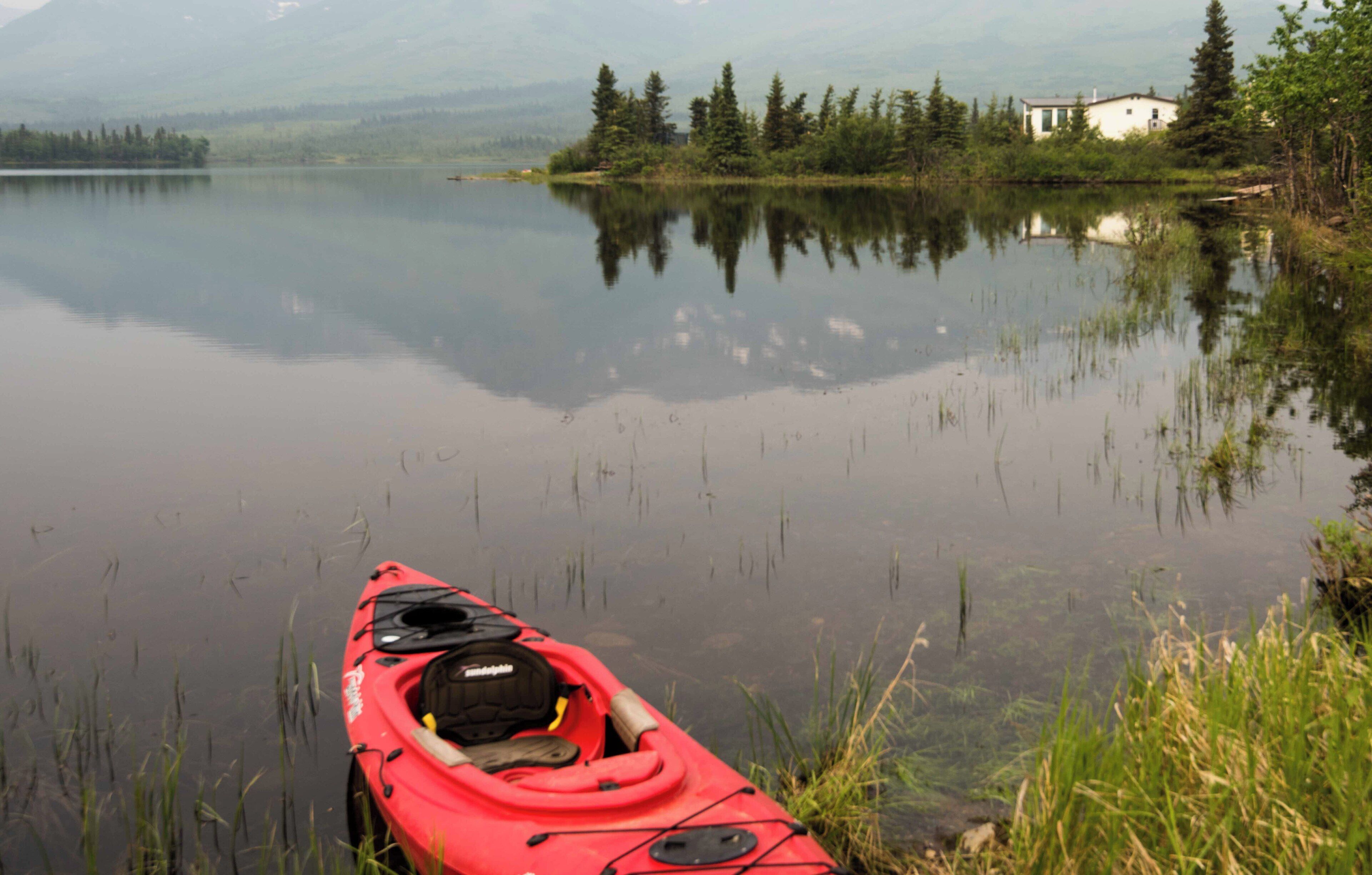 A red kayak on calm water with a house and trees in the background.