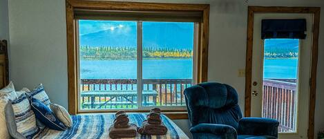 A room with a large window offering a view of a lake and dock. There is a bed with striped bedding and a blue chair.