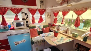 Una cocina compacta y bien organizada con microondas, tetera y varios utensilios de cocina. Hay una pequeña zona de comedor con una mesa y sillas, y una ventana con cortinas rojas. La habitación tiene un suelo a cuadros y una cama con ropa de cama de lunares rojos y blancos.