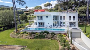Una casa moderna de dos pisos con piscina, rodeada de abundante vegetación y colinas al fondo.