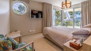Un dormitorio con una cama grande, un televisor montado en la pared, un ventanal redondo y un balcón con vistas a árboles.