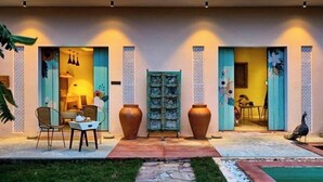 A modern building with a stone pathway leading to a door, a small outdoor seating area with chairs and a table, and a bird on the grass. - Parijat Private Pool Villa 1, 2 and 3 BHK (Udaipur)