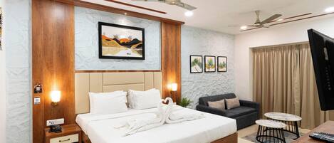 A modern hotel room with a large bed, a sofa, two small tables, ceiling fans, and a TV.