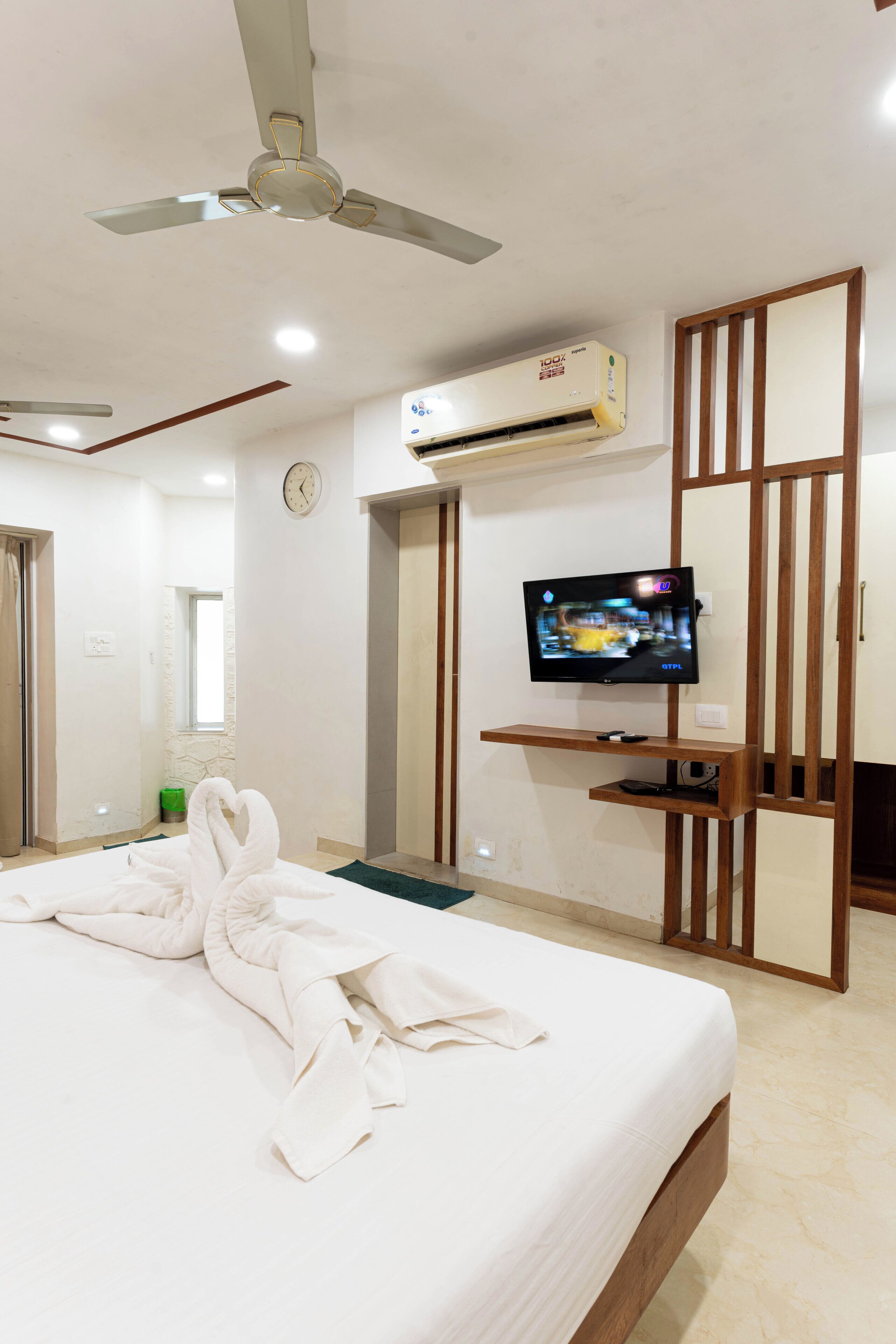 Una camera d'albergo con un letto, una ventola a soffitto, un condizionatore d'aria, una televisione e un pannello divisorio in legno.