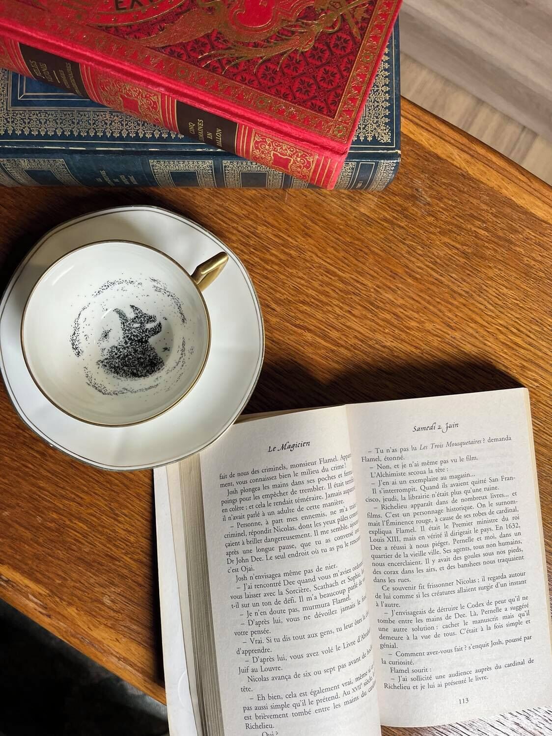 A cup of coffee on a saucer, a book titled "Le Magicien" on a wooden table, and a red book with gold designs in the background.