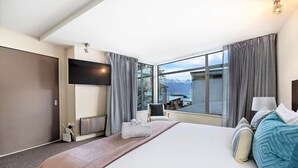 A modern hotel room with a large bed, a flat-screen TV mounted on the wall, a view of mountains through the window, and a small table with chairs. - Manaaki Alpine Retreat Central CBD ~ SPA ~ Garage (Queenstown)