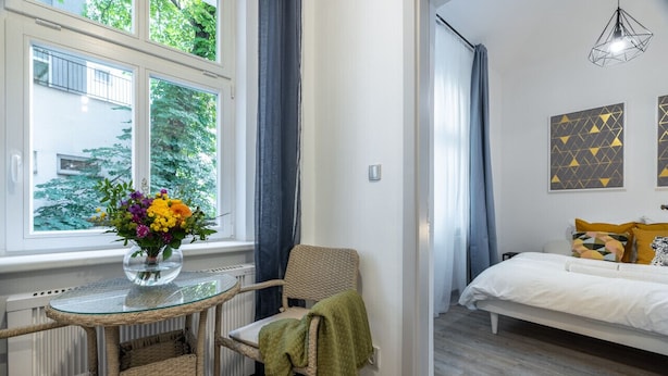 Une pièce avec une table en verre, des chaises en osier, un lit avec une literie blanche et une fenêtre avec des rideaux.