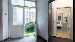 Un couloir avec une porte vitrée donnant sur un balcon, une chambre avec un lit et une table de chevet, et une vue sur les bâtiments à l’extérieur.