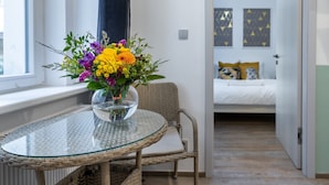 Une pièce avec une table à plateau en verre, une chaise en osier et un lit recouvert d’une couette blanche.