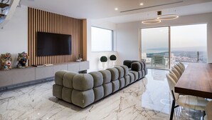 Ein modernes Wohnzimmer mit großem Fernseher, einer Eckcouch, einer Essecke mit Holztisch und Blick auf die Stadt durch große Fenster.
