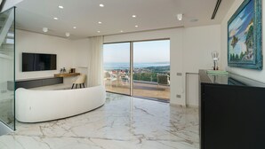 Ein modernes Wohnzimmer mit einer gebogenen weißen Couch, einem Flachbildfernseher, einem Essbereich mit Tisch und Stühlen sowie einem großen Fenster mit Blick auf die Stadt.