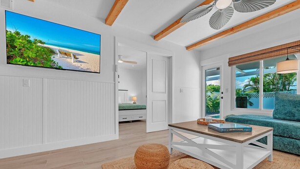 Uma sala de estar moderna com televisão de tela plana, mesa de centro de madeira, um banquinho trançado e vista para a praia através de grandes janelas.