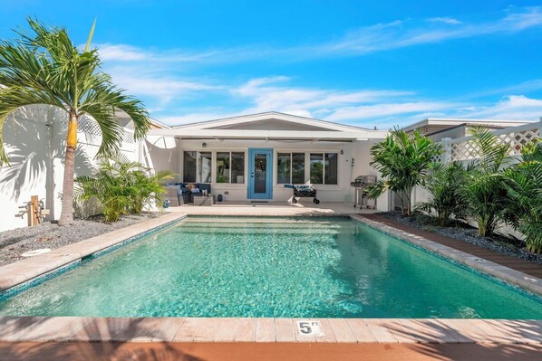 Ein Pool mit klarem, blauem Wasser, ein weißes Gebäude mit blauer Tür und eine Terrasse mit Gartenmöbeln.
