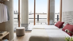 Ein modernes Hotelzimmer mit großem Fenster und Blick auf ein Gewässer, einem Bett mit roten Kissen, einem kleinen runden Hocker und einem Holzregal mit Büchern.