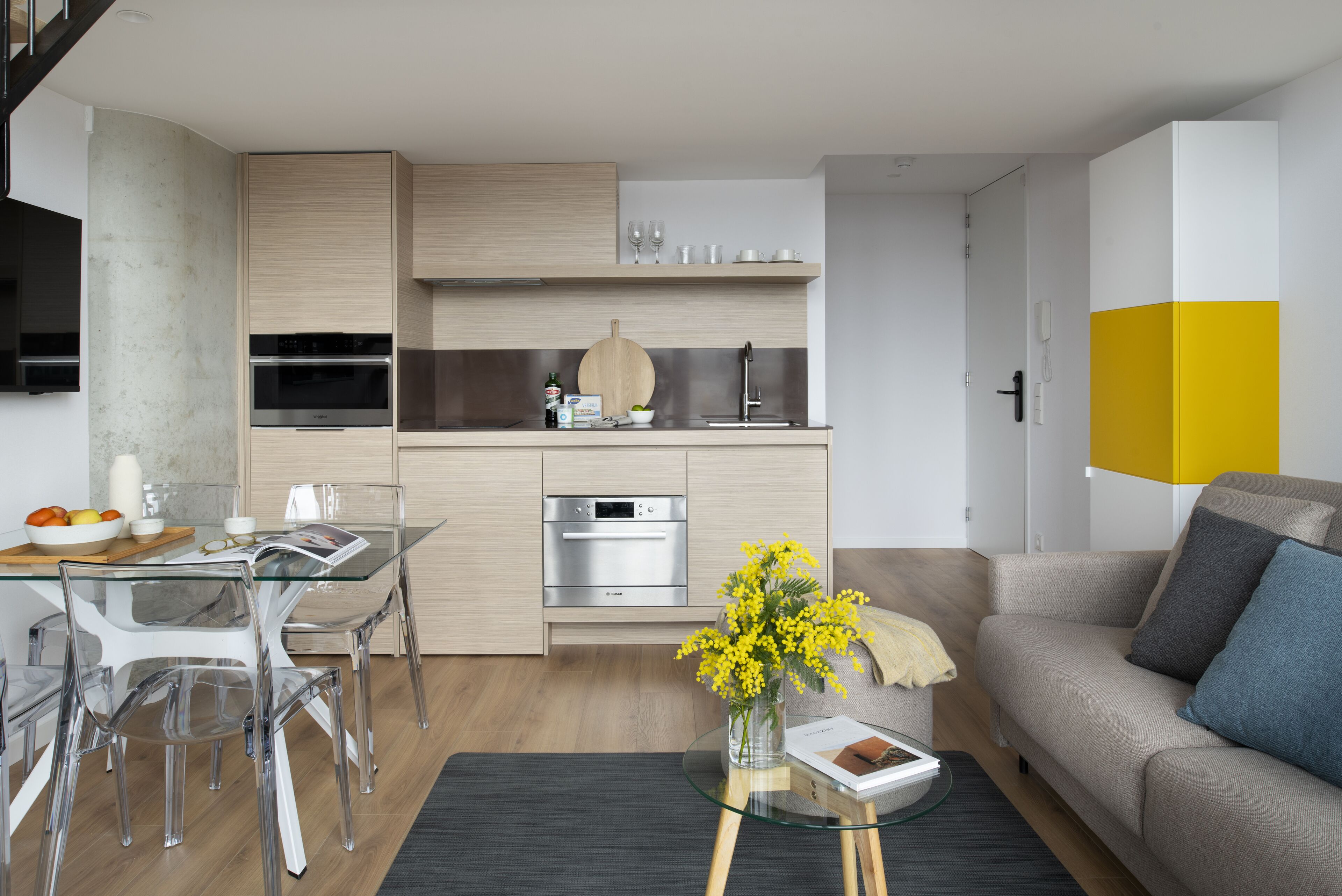Una cucina moderna con elettrodomestici integrati, una zona pranzo con sedie trasparenti e un soggiorno con divano e tavolino.