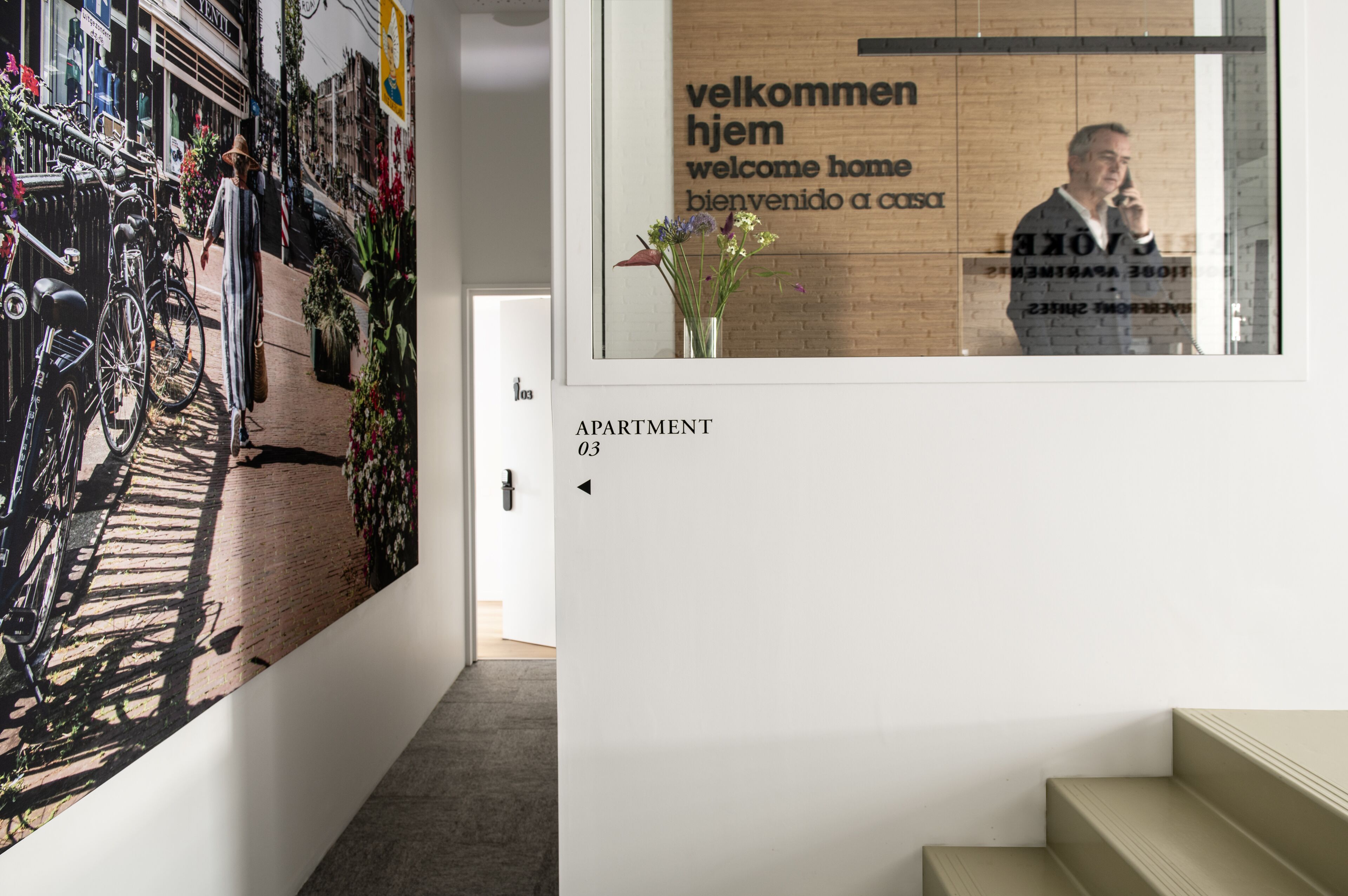 A hallway with a bicycle rack, a welcome sign in multiple languages, and an apartment number sign.