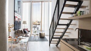 Ein modernes Wohnzimmer mit einem Glastisch, einer Couch und einem Essbereich mit Stühlen. Eine Treppe mit schwarzer Handlauf führt auf eine erhöhte Ebene.