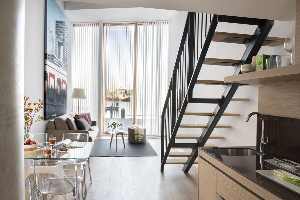 Ein modernes Wohnzimmer mit einem Glastisch, einer Couch und einem Essbereich mit Stühlen. Eine Treppe mit schwarzer Handlauf führt auf eine erhöhte Ebene.