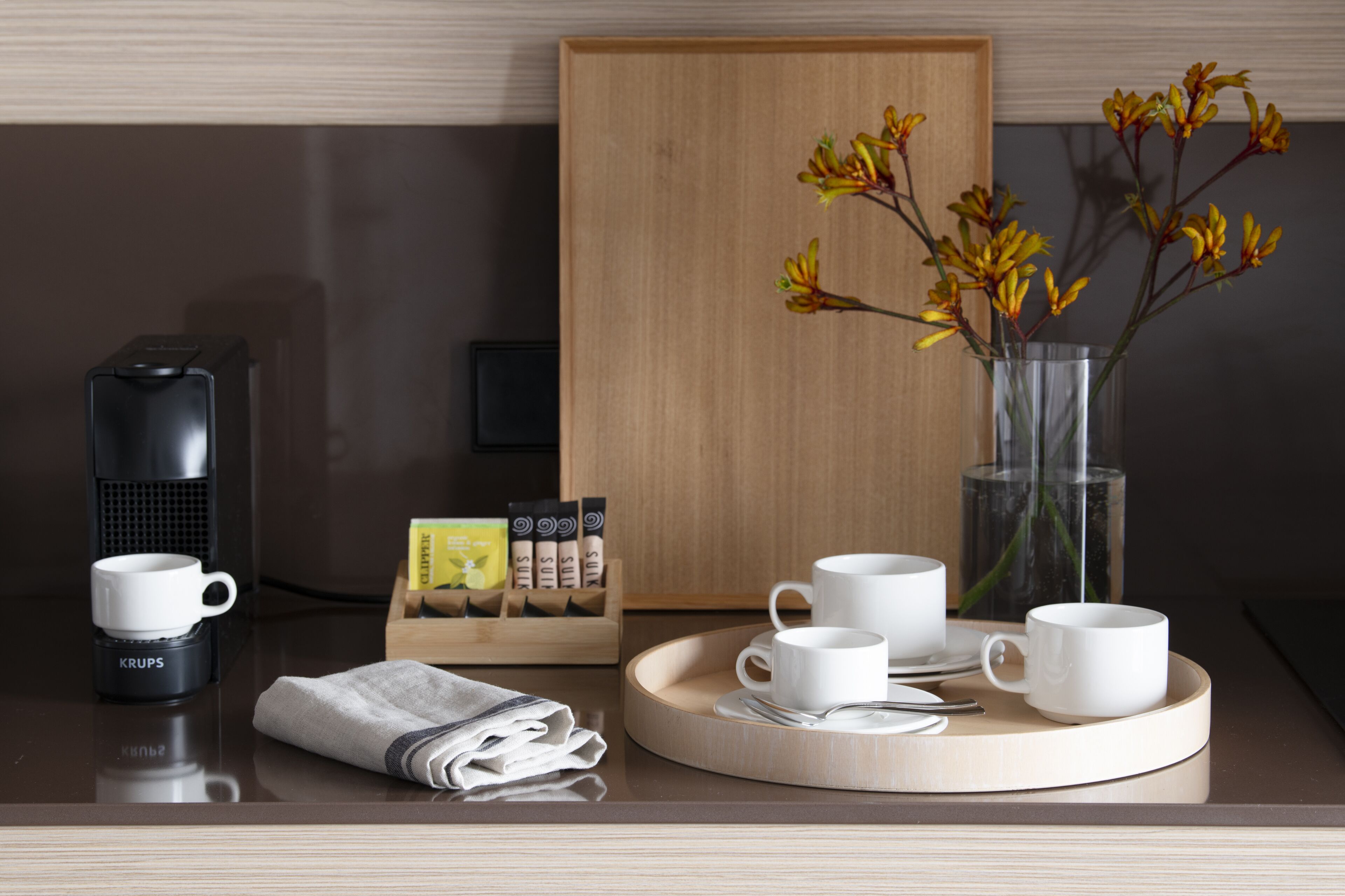 Een keurig opgemaakte ontbijtsetting met een koffiezetapparaat, kopjes, een dienblad en een vaas met bloemen.