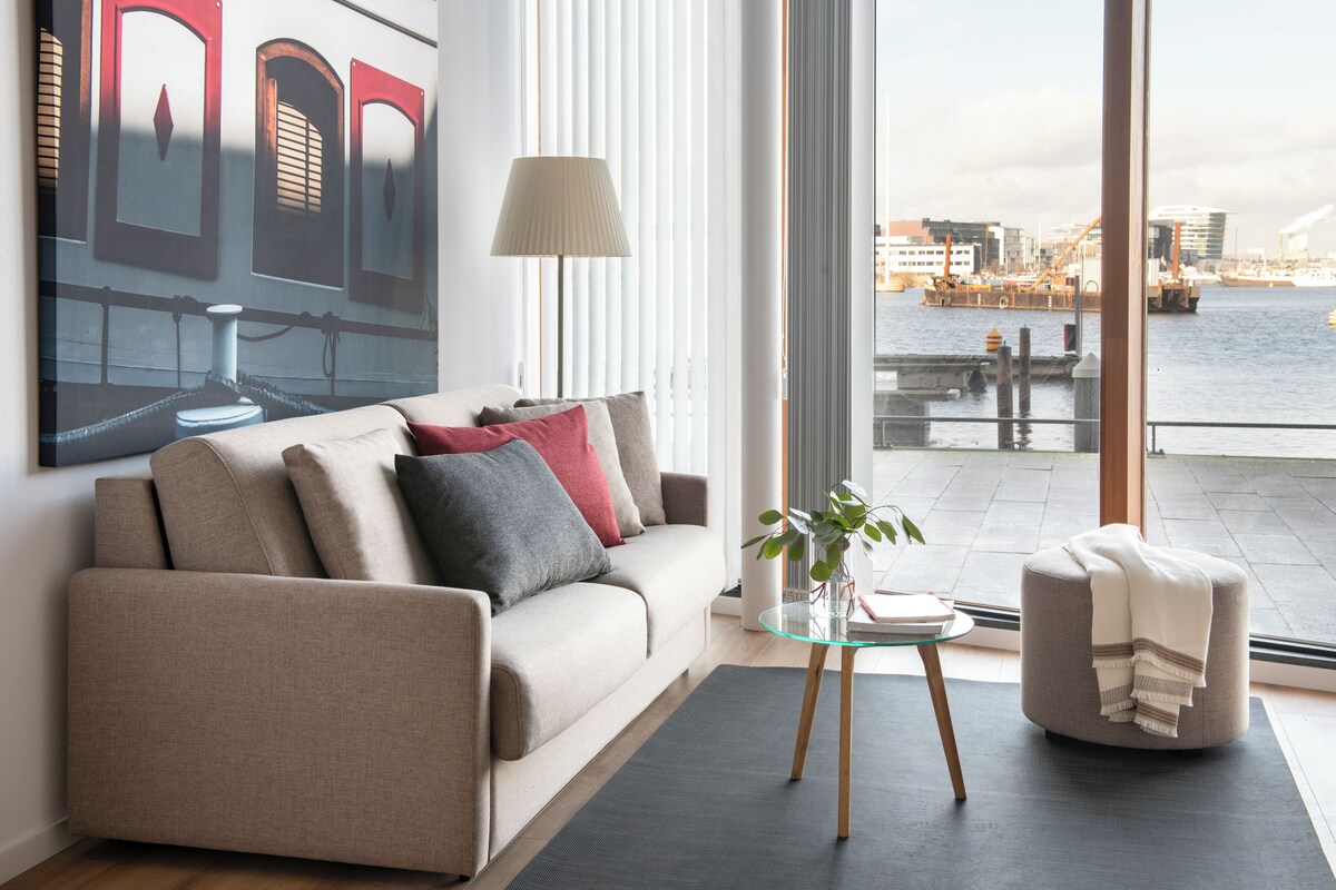 A modern living room with a sofa, a glass coffee table, and a view of the water and cityscape.