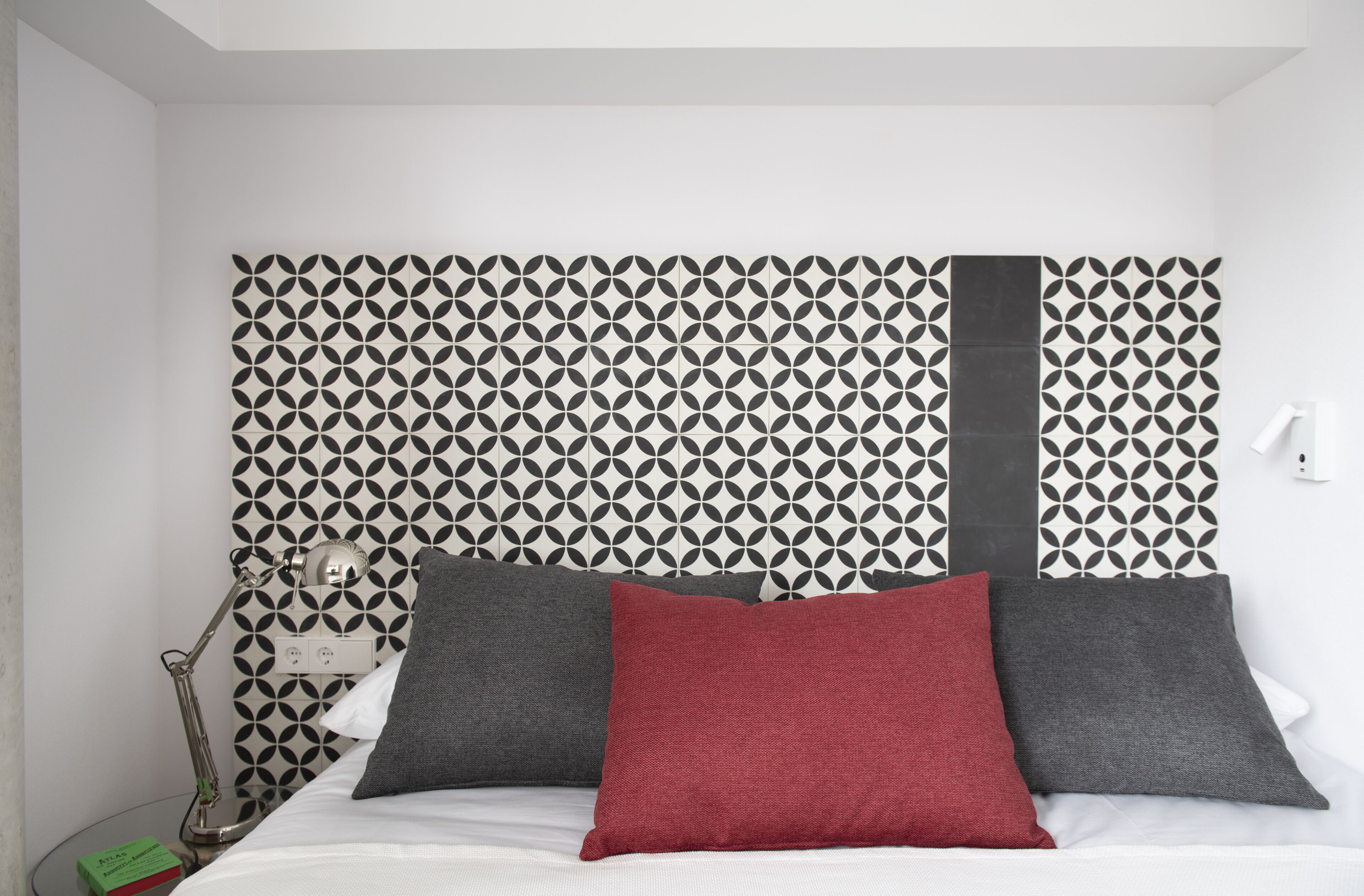 A bed with a patterned headboard, two pillows, a bedside lamp, and a green book on a bedside table.