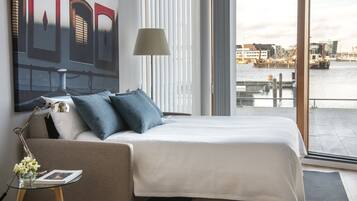 A hotel room with a bed, a lamp, a glass table with a book and flowers, and a large window with a city view.
