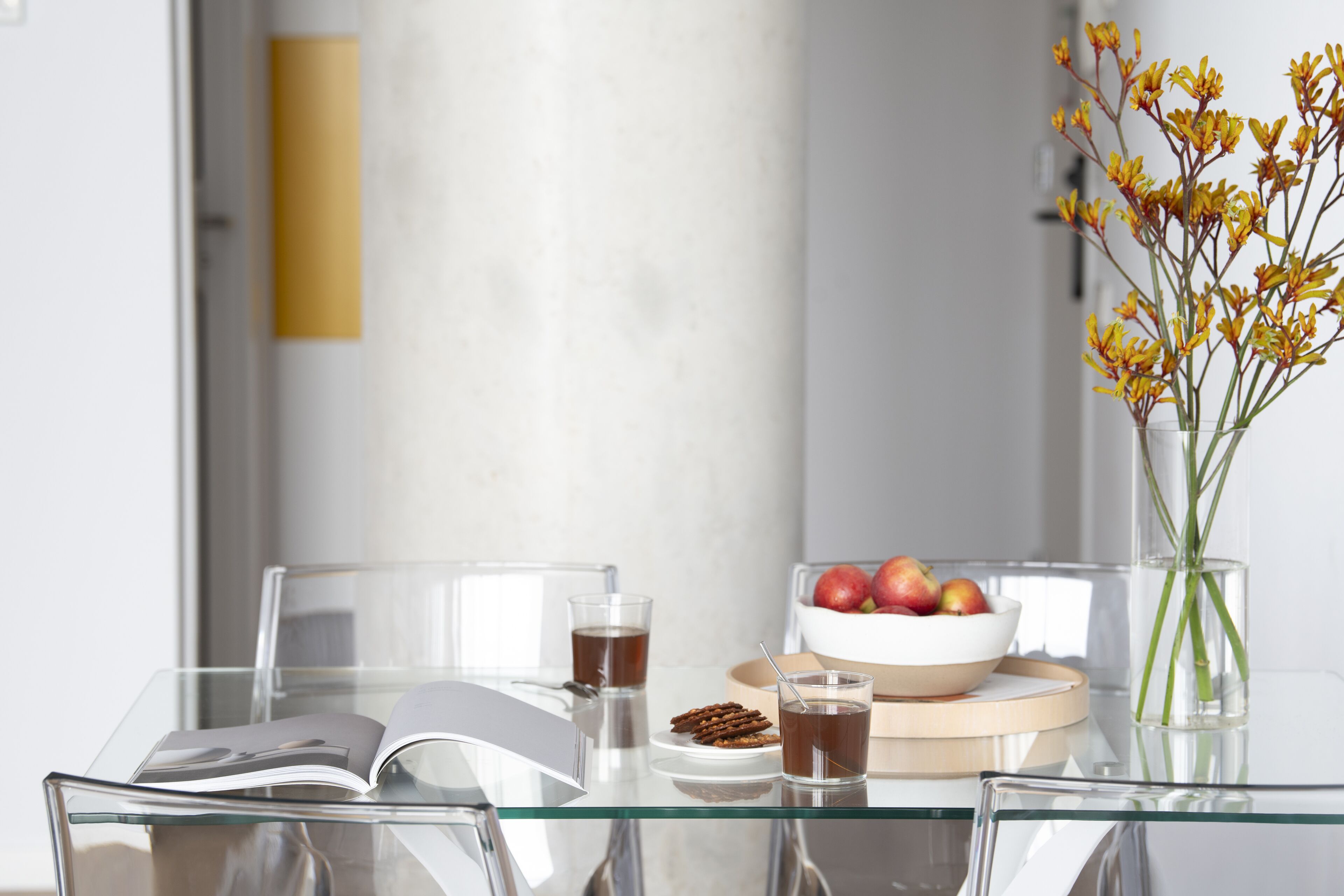 A modern dining table with a glass top, a bowl of fruit, a glass of tea, and a book.