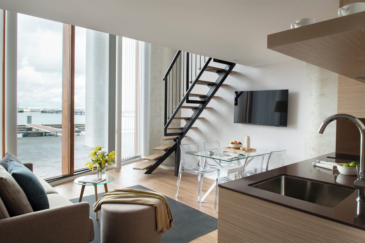 A modern kitchen with a large island, a dining area with clear glass chairs, a staircase, and a view of a body of water through large windows.