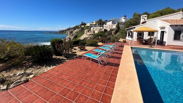 Zona de piscina con vistas despejadas al océano, patio empedrado y edificio con fachada blanca y tejado rojo.